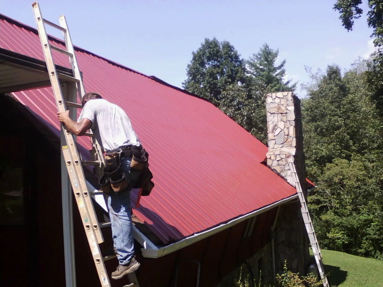 Roofer on a ladder beside a red metal roof and stone chimney — Soffit & Fascia Repair by Roofing Brothers in Welton